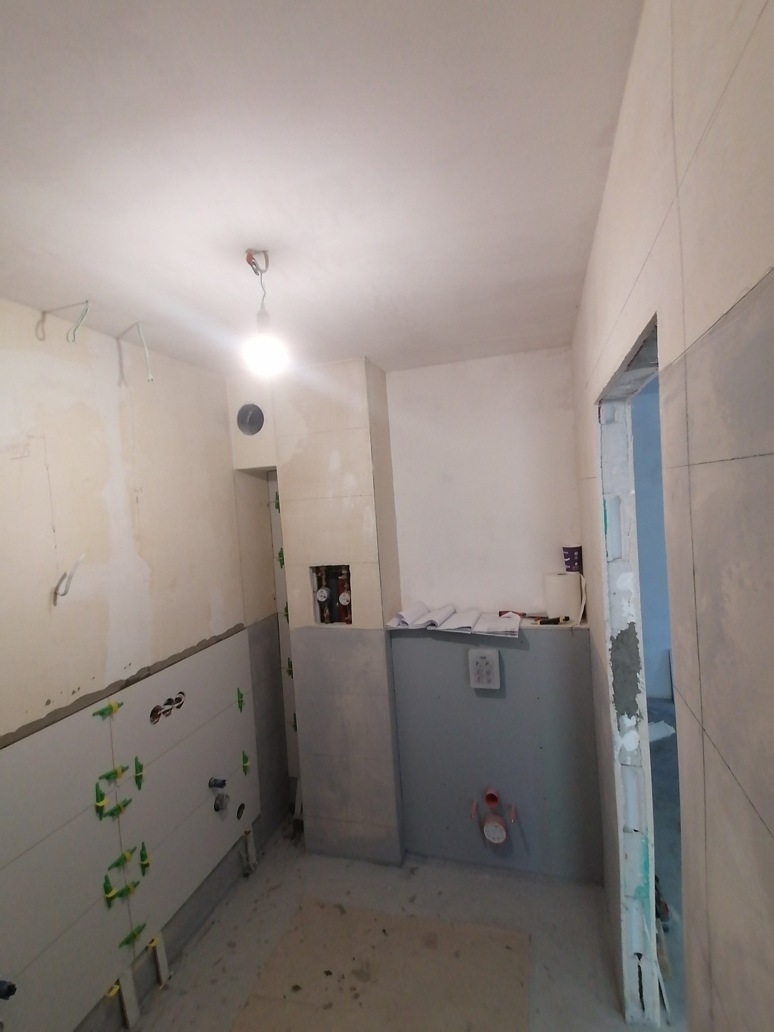 Łazienka w trakcie remontu: częściowo ułożone jasne płytki na ścianie z zielonymi krzyżykami dystansowymi, widoczne instalacje hydrauliczne, przygotowane ściany do dalszych prac wykończeniowych.