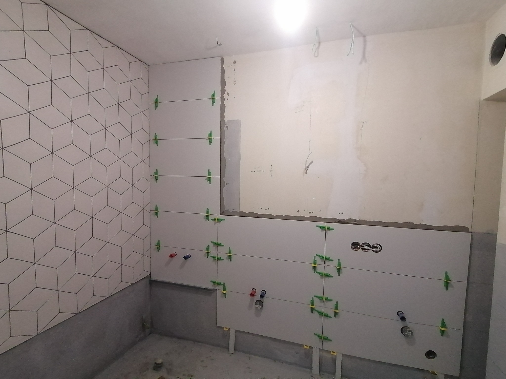 Ściana łazienki w trakcie układania płytek z geometrycznym wzorem, widoczne krzyżyki dystansowe, rury doprowadzające wodę i otwory na gniazdka elektryczne.