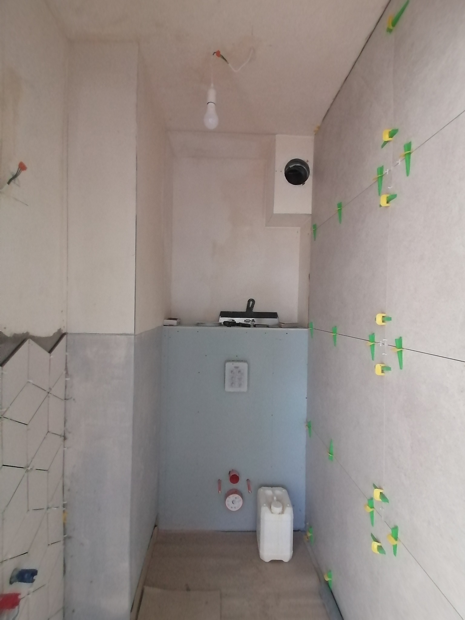 Wnętrze łazienki w trakcie remontu: częściowo ułożone płytki na ścianach, z użyciem poziomicy i klipsów dystansowych, widoczna instalacja wodna i kanister, surowa ściana w tle.