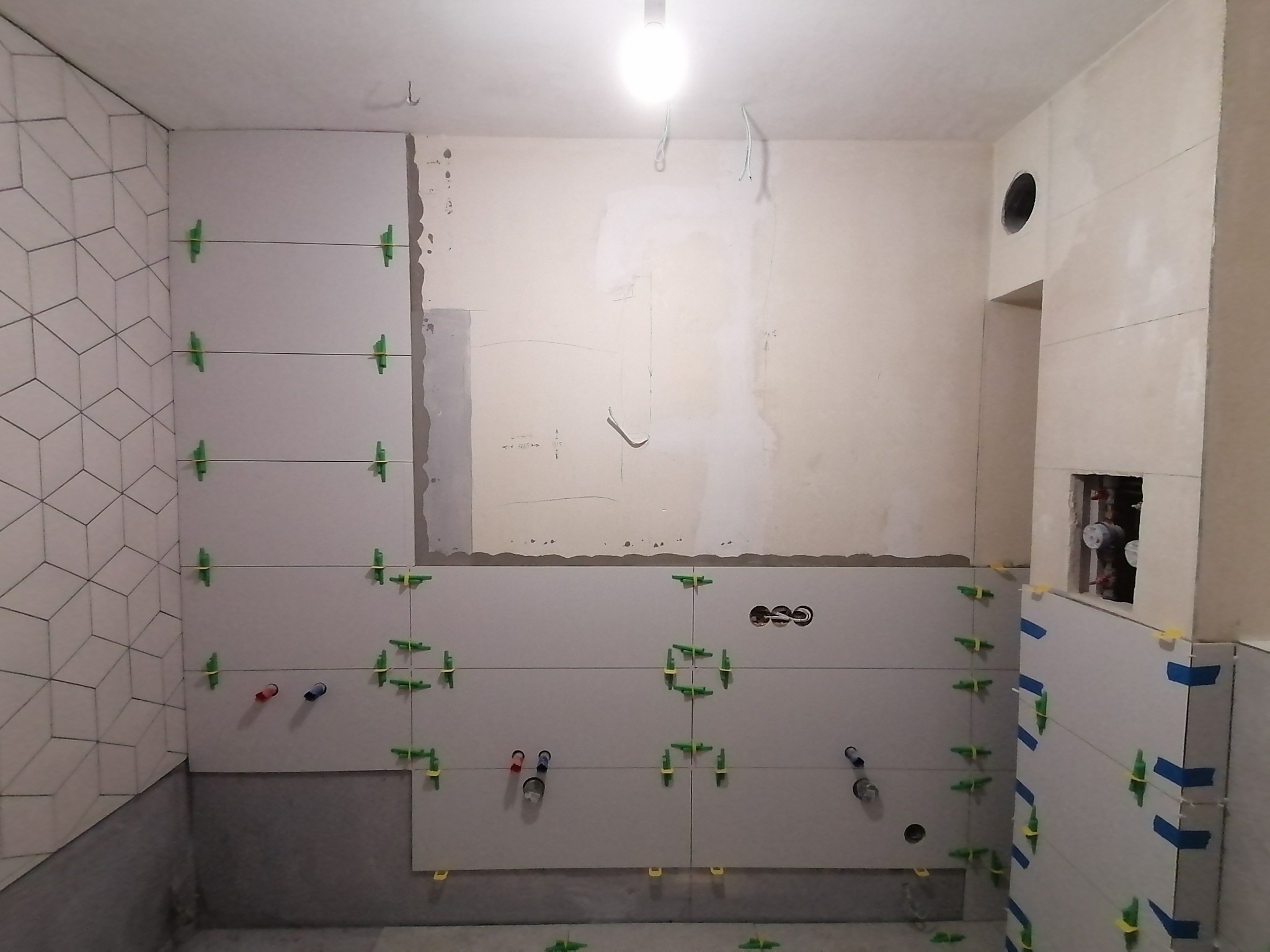 Ściana w trakcie układania płytek ceramicznych w łazience, widoczne krzyżyki dystansowe, fragment ściany z wzorem geometrycznym i przygotowane podejścia do instalacji wodnej.
