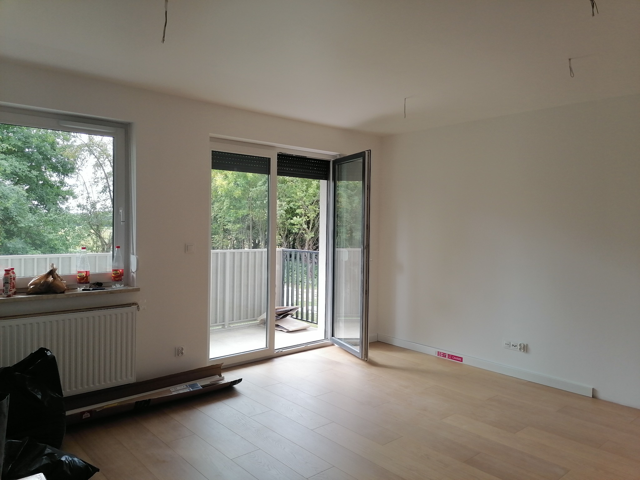 Puste, jasne pomieszczenie z parkietem, otwartym balkonem i oknem, widoczne kable zwisające z sufitu i karton z listwami przypodłogowymi przy ścianie.