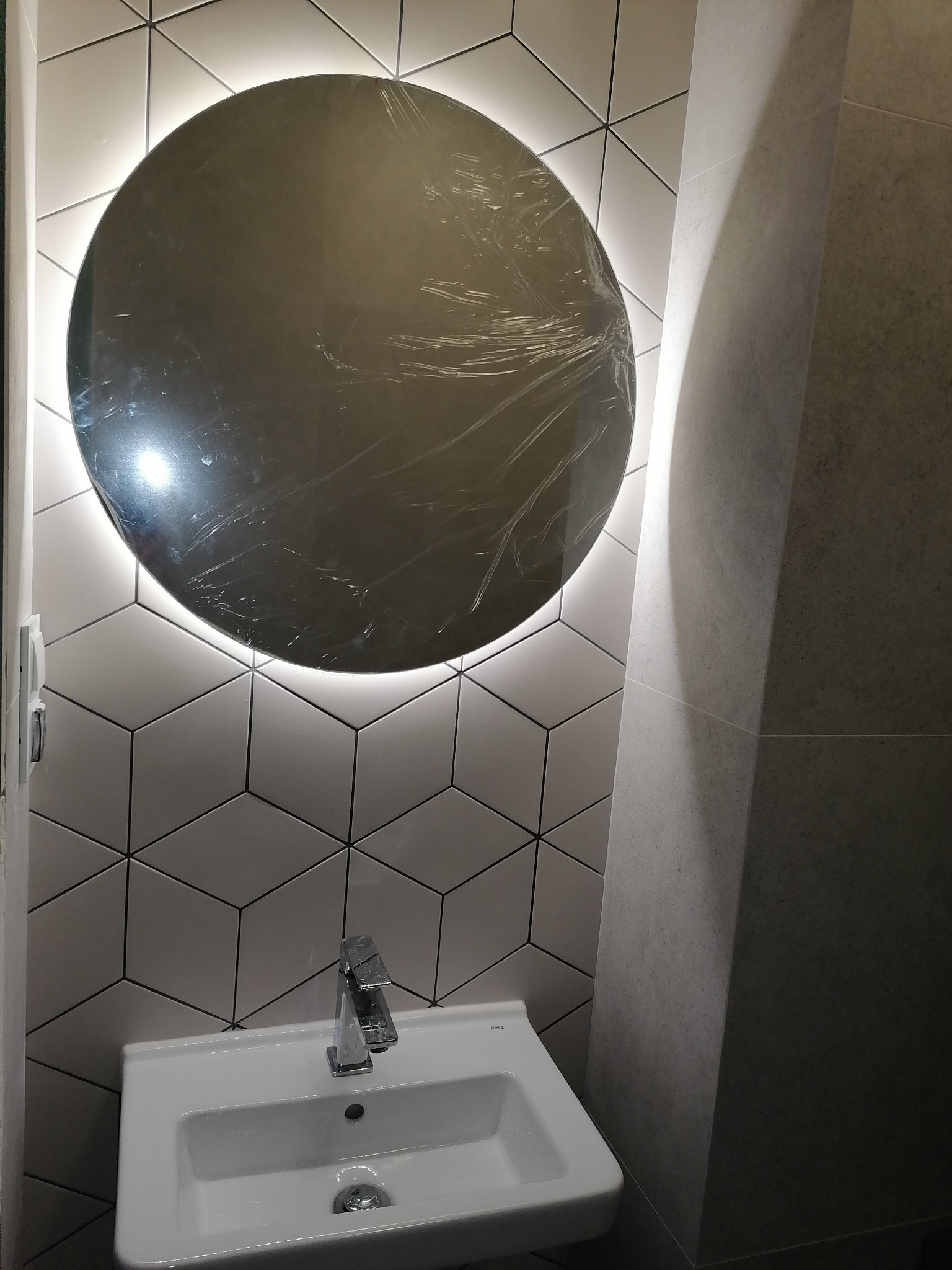 Nowoczesna łazienka z okrągłym lustrem podświetlanym LED, umywalką z chromowaną baterią i heksagonalnymi płytkami ściennymi, lustro jeszcze w folii ochronnej.