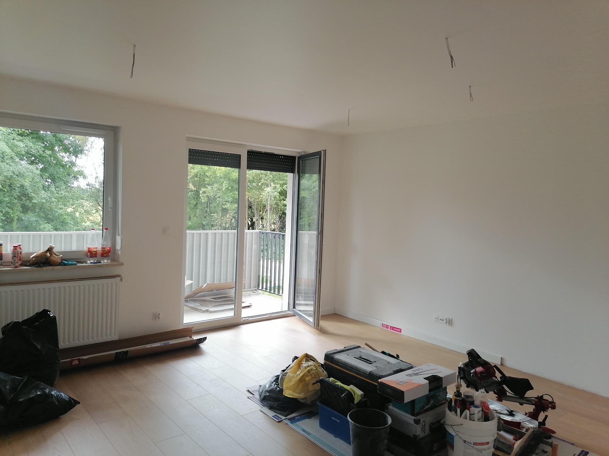 Pusty pokój z otwartym balkonem i narzędziami przygotowanymi do remontu, widoczne kable wystające z sufitu, panele podłogowe i skrzynki narzędziowe.