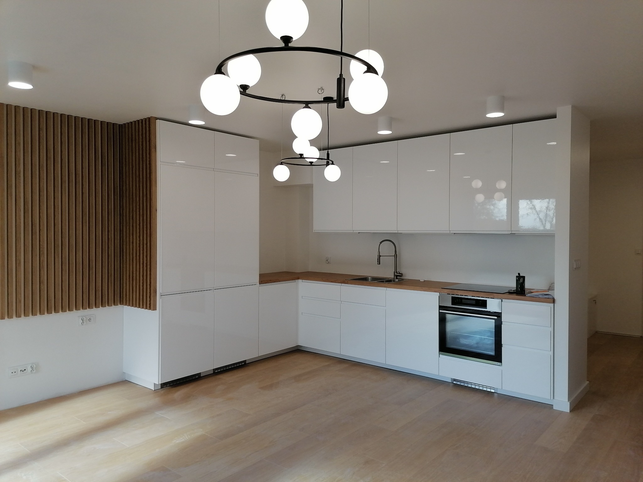 Nowoczesna kuchnia z białymi, lakierowanymi szafkami, drewnianym blatem i podłogą, oświetlona okrągłym żyrandolem z kulistymi kloszami i drewnianą ścianką dekoracyjną z pionowych listew.