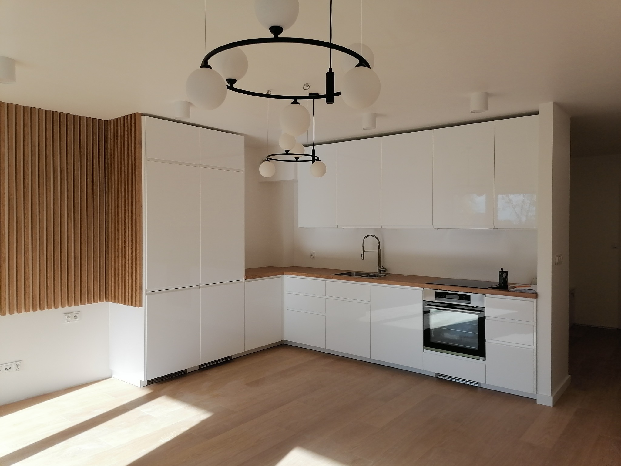 Nowoczesna, minimalistyczna kuchnia z białymi, lakierowanymi szafkami, drewnianym blatem i dekoracyjną ścianą z drewnianych listewek. Okrągła lampa sufitowa z białymi kulami nad wyspą kuchenną.