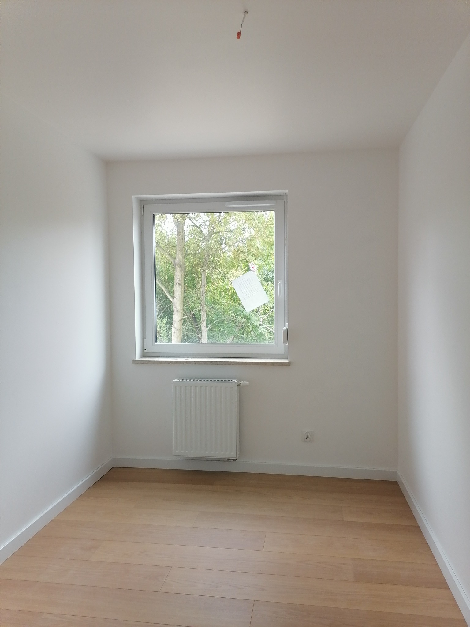 Pusty pokój po remoncie: świeżo pomalowane białe ściany, jasna drewniana podłoga, białe listwy przypodłogowe, okno z widokiem na drzewa i biały grzejnik pod nim.