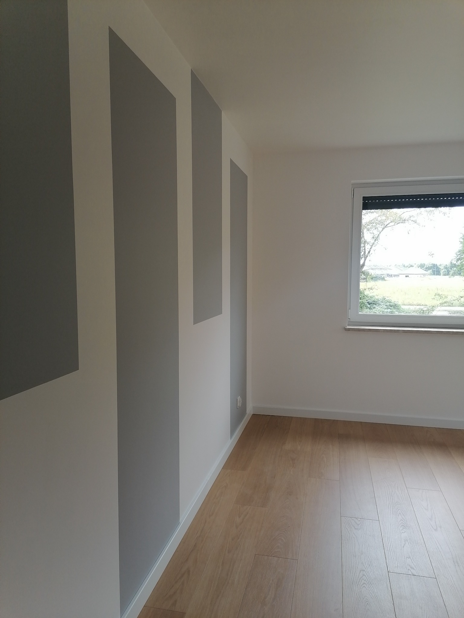 Wnętrze pokoju po malowaniu ścian w pionowe pasy szarości i bieli, z drewnianą podłogą i oknem z widokiem na zieleń.
