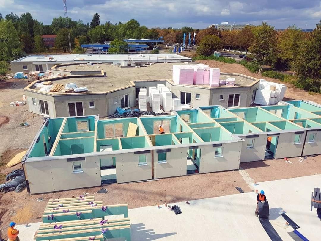 Budowa modułowego budynku z prefabrykatów, widoczne elementy konstrukcyjne i pracownicy w kaskach.