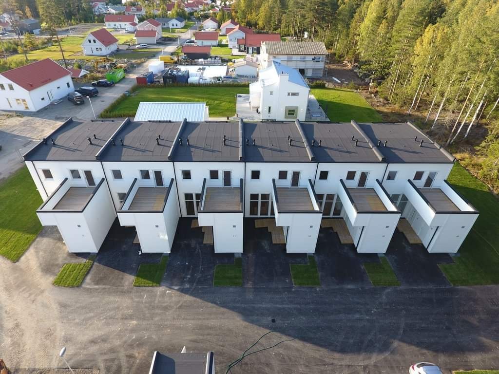 Widok z lotu ptaka na szereg nowych, bliźniaczych domów z płaskimi, ciemnymi dachami i białymi elewacjami, otoczonych zielenią i innymi budynkami mieszkalnymi.