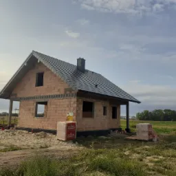 Stan surowy otwarty domu z czerwonej cegły w Sarbinowie, widoczne palety z materiałem budowlanym Porotherm oraz czarna dachówka.