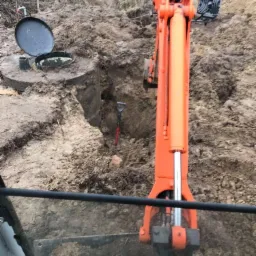 Pomarańczowa koparka w trakcie prac ziemnych przy betonowym zbiorniku z otwartym włazem, widok z kabiny operatora.