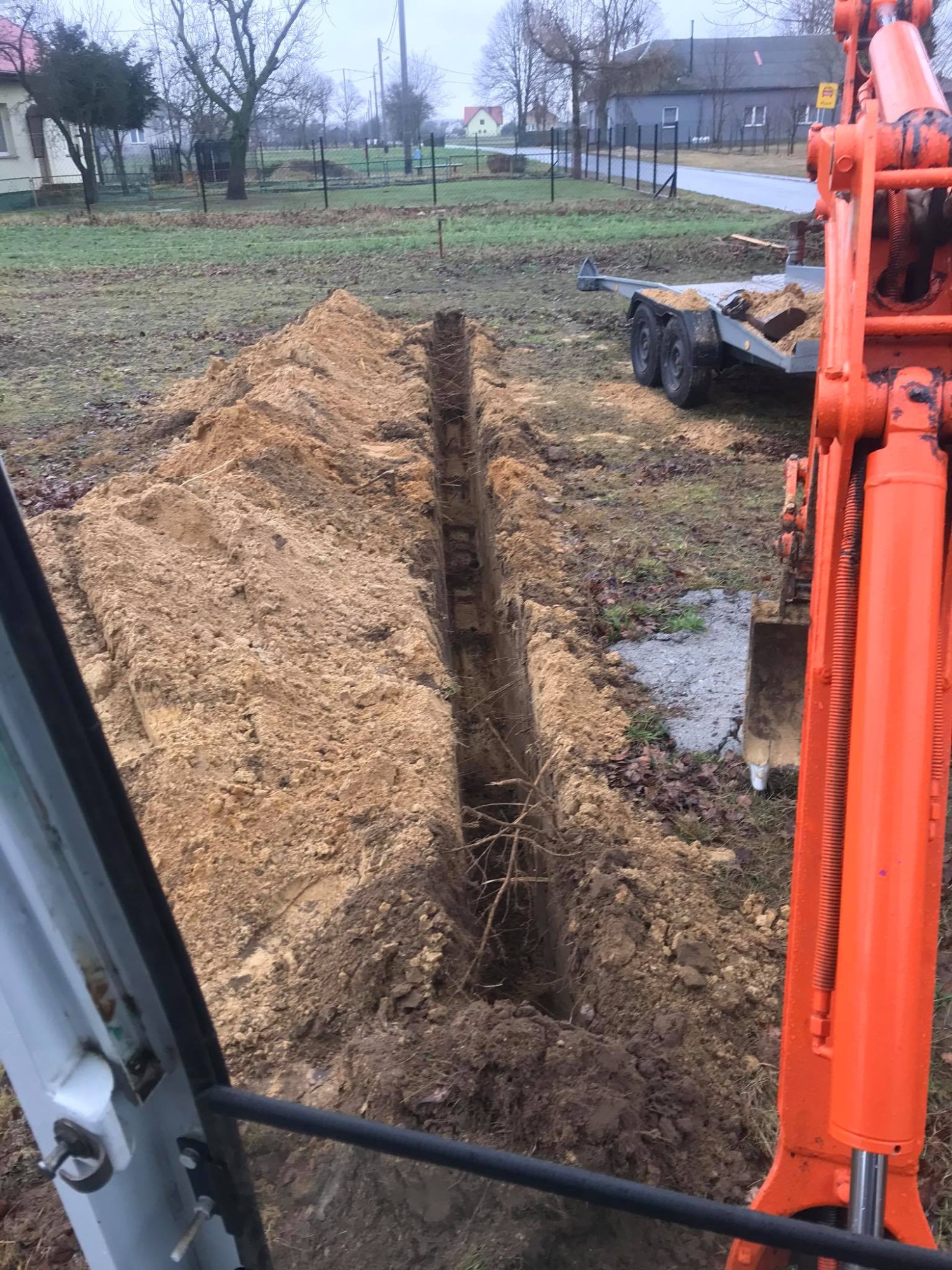 Wąski wykop liniowy w ziemi, widoczny z kabiny koparki, z pomarańczowym ramieniem koparki po prawej stronie i przyczepą w tle.