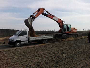 Pomarańczowa koparka nabiera ziemię i wysypuje ją na białą platformę ciężarówki na tle pola i drzew.