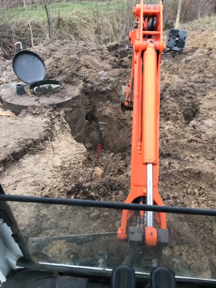 Pomarańczowa koparka w trakcie prac ziemnych przy betonowym zbiorniku z otwartym włazem, widok z kabiny operatora.