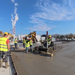 ZHUP MIRS - Wylewanie betonu na dużej powierzchni przez ekipę budowlaną z użyciem specjalistycznej maszyny. W tle kominy i błękitne niebo.