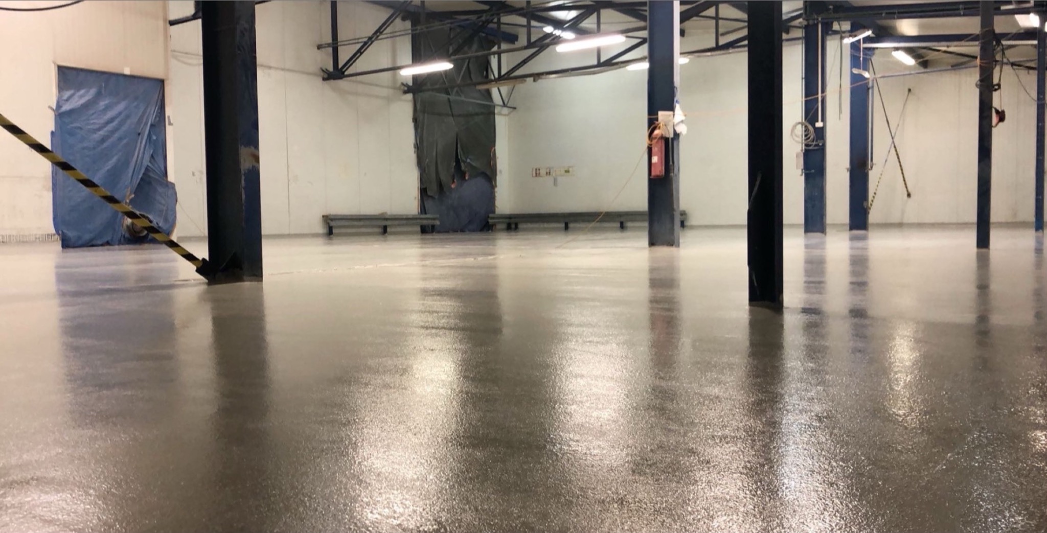 Wnętrze hali po aplikacji izolacji natryskowej w Bełchatowie. Widoczne słupy konstrukcyjne i gładka, błyszcząca powierzchnia podłogi. Oświetlenie sufitowe.