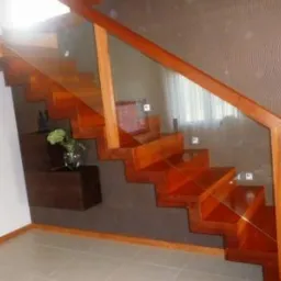 Nowoczesne schody z drewna egzotycznego, z przeszkloną balustradą, zamontowane na ścianie z dekoracyjną tapetą. Pod schodami wisząca szafka z dekoracją roślinną.