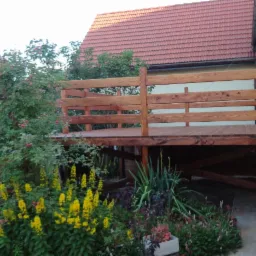 Drewniany taras z prostą, poziomą balustradą, otoczony kwitnącą roślinnością, z widoczną konstrukcją nośną i schodami prowadzącymi na trawnik.