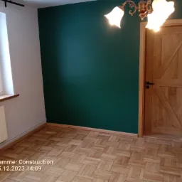 Wnętrze pokoju z parkietem w jodełkę, pomalowaną na zielono ścianą i drewnianymi drzwiami w stylu rustykalnym pod oświetleniem klasycznego żyrandola.