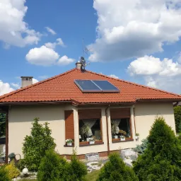 Dom jednorodzinny z czerwoną dachówką, na której zamontowane są dwa panele fotowoltaiczne; na dachu widoczna antena satelitarna i komin; wokół domu zadbany ogród z licznymi krzewami.
