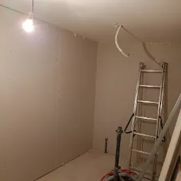 Wnętrze pokoju w trakcie remontu, ściany wyłożone płytami gipsowo-kartonowymi, widoczne ślady po wkrętach, drabina oparta o ścianę, na podłodze odkurzacz przemysłowy i resztki materiałów...