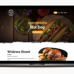 Ekran laptopa wyświetlający stronę internetową restauracji z sliderem prezentującym zdjęcia hot dogów i burgerów, widoczne logo w lewym górnym rogu i menu w prawym górnym rogu.