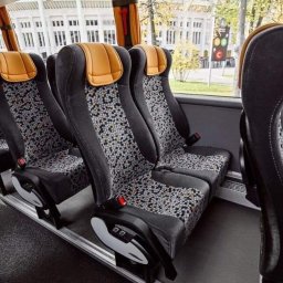 LUX-BUS przewóz osób - Wnętrze autobusu turystycznego z rzędami czarno-szarych siedzeń z pomarańczowymi zagłówkami i wzorzystą tapicerką, widok z perspektywy pasażera, za oknem budynek i sygnalizacja świetlna.