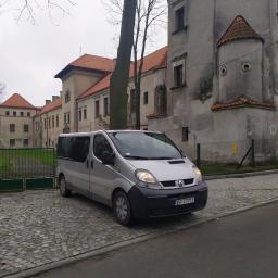 LUX-BUS przewóz osób - Srebrny van Renault Trafic zaparkowany na brukowanej nawierzchni przed zabytkowym budynkiem z czerwonym dachem i wieżyczką.