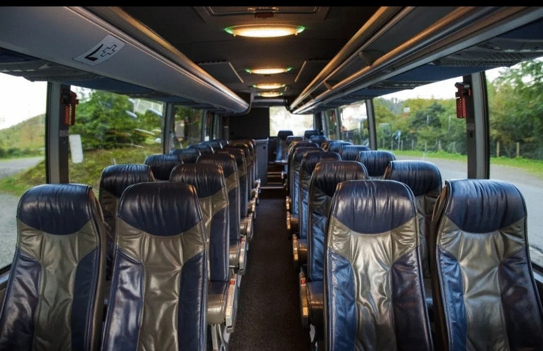 Wnętrze autobusu turystycznego z rzędami ciemnych, skórzanych foteli z szarymi wstawkami, widok z przodu na puste przejście i okna z widokiem na zieleń na zewnątrz.
