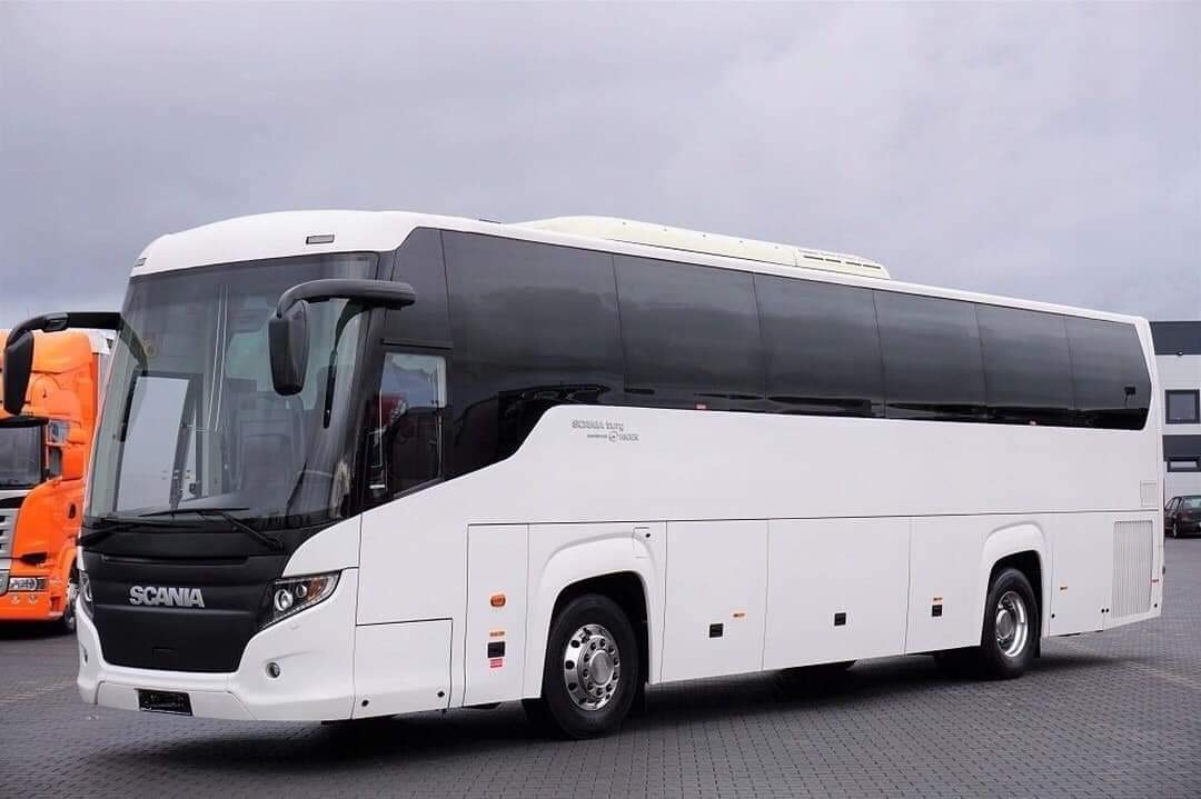 Biały autokar marki Scania z przyciemnianymi szybami, zaparkowany na placu z szarą kostką brukową, z pomarańczową ciężarówką w tle po lewej stronie.