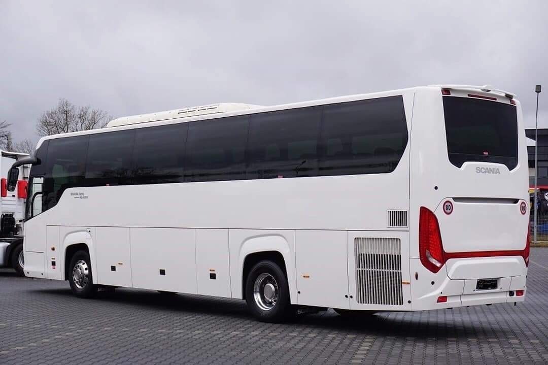 Biały autokar marki Scania z przyciemnianymi szybami, widok z tyłu na parkingu z kostki brukowej, obok zaparkowany ciągnik siodłowy.