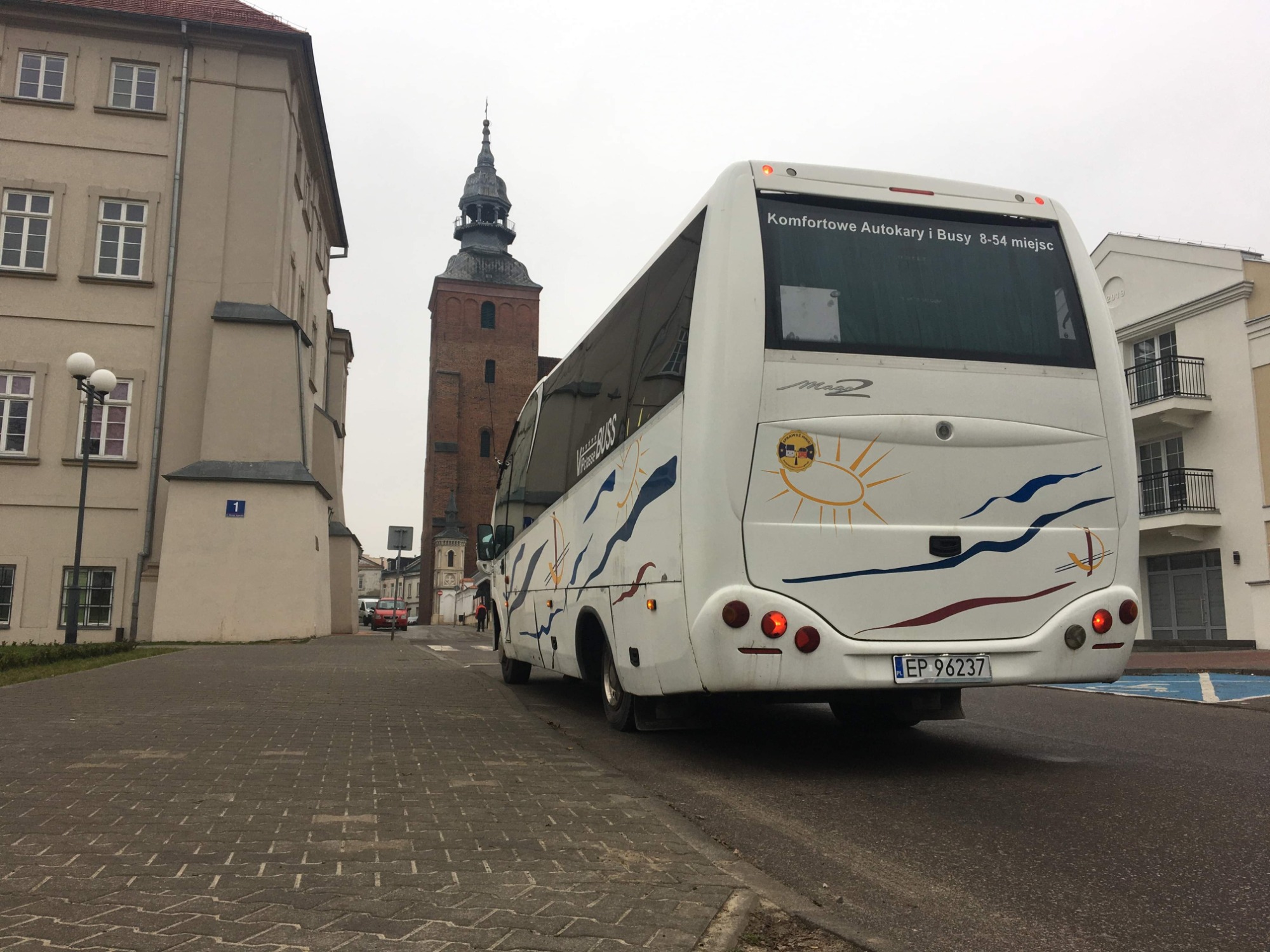 Biały autokar z grafiką słońca i fal, zaparkowany na ulicy z brukowaną kostką, w tle budynek z wieżą.