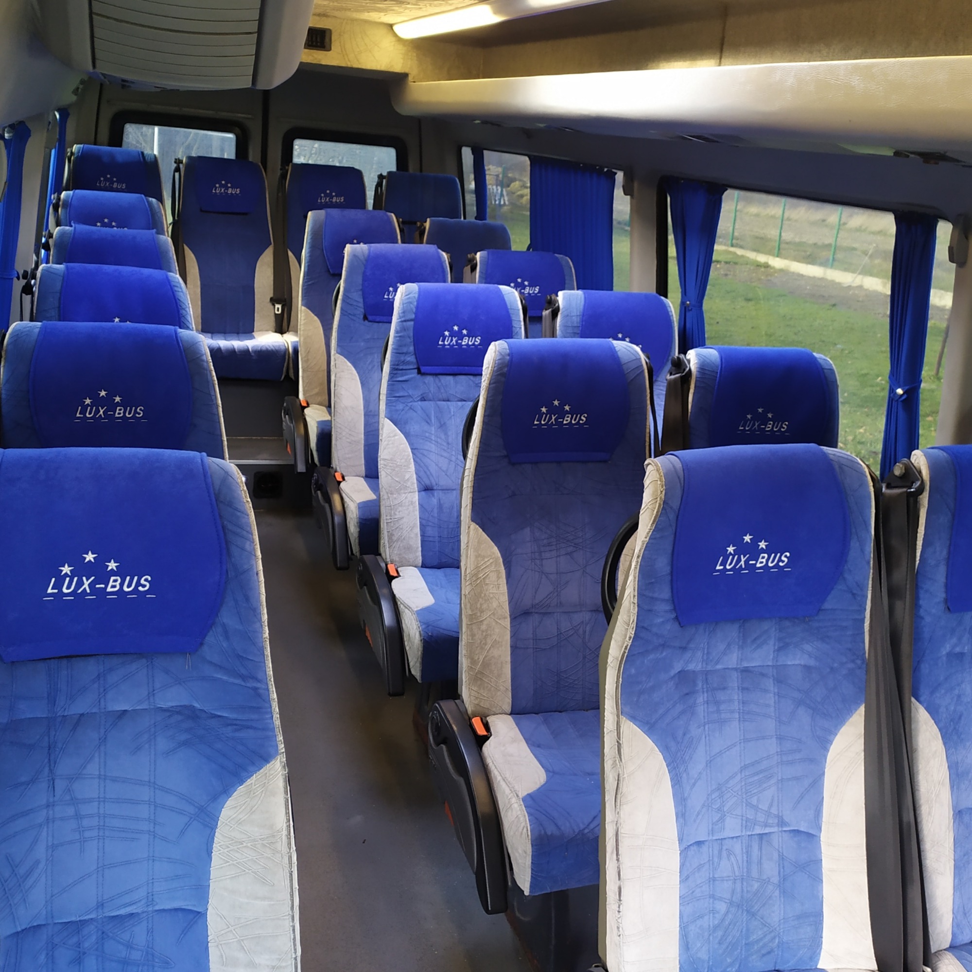 Wnętrze autobusu z rzędami niebiesko-szarych foteli z logo 'LUX-BUS' na zagłówkach, widok z przodu pojazdu w kierunku okna z zasłoną.