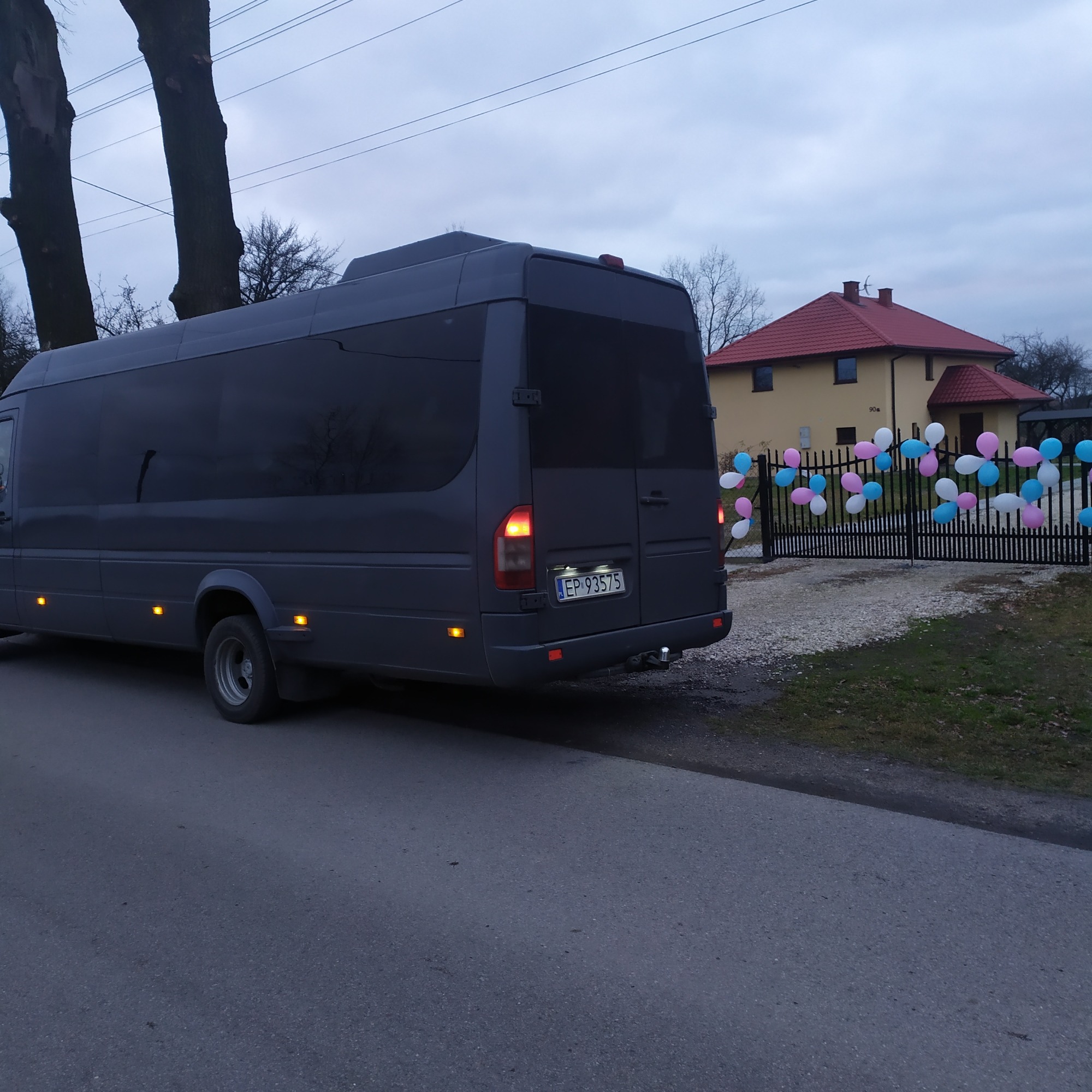 Szary bus zaparkowany na ulicy przed ogrodzeniem domu udekorowanym balonami w kolorach różowym, białym i niebieskim.