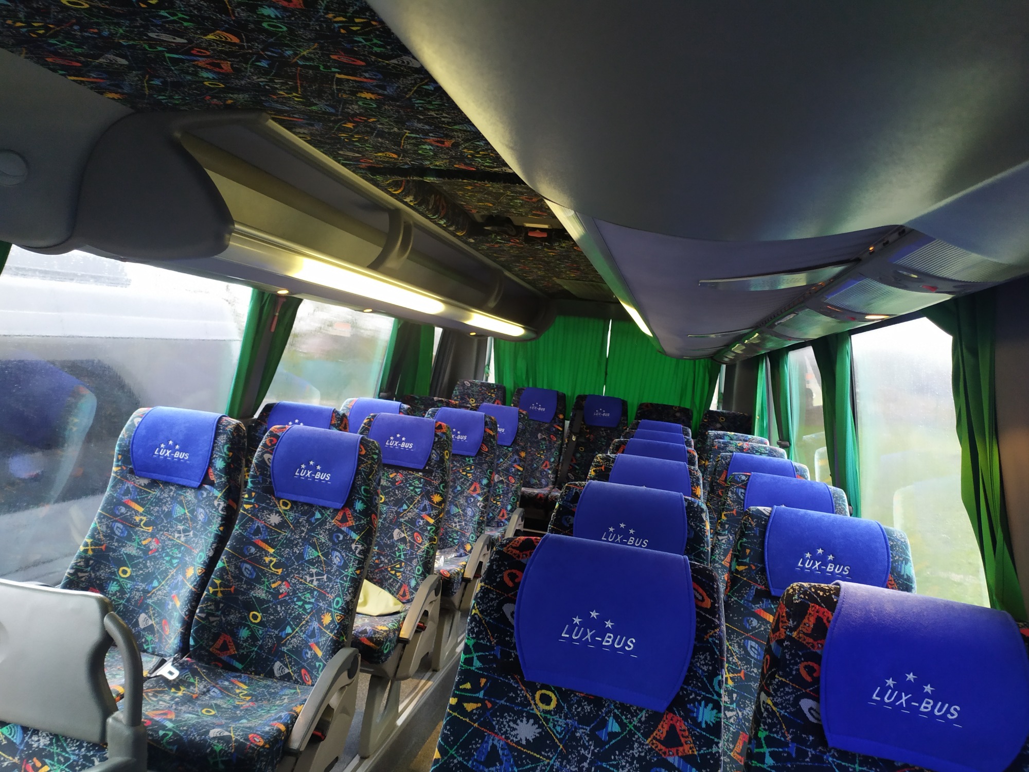 Wnętrze autobusu dalekobieżnego z rzędami foteli obitych wzorzystą tkaniną i niebieskimi zagłówkami z logo firmy przewozowej, widok z perspektywy pasażera.
