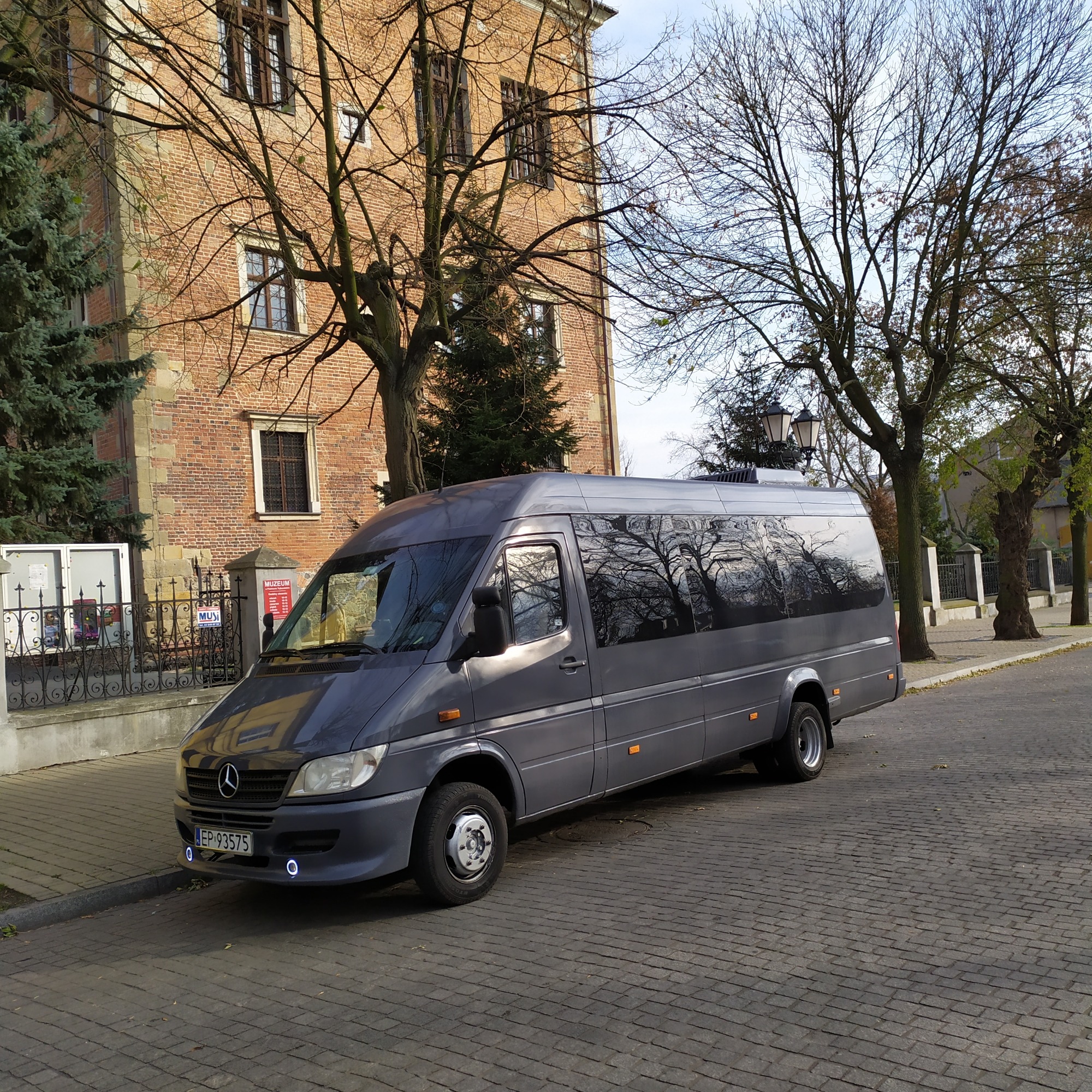Szary bus Mercedes Sprinter zaparkowany na brukowanej ulicy, z widocznym budynkiem z czerwonej cegły w tle i drzewami z nagimi gałęziami odbijającymi się w oknach, z niebieskimi światłami LED...