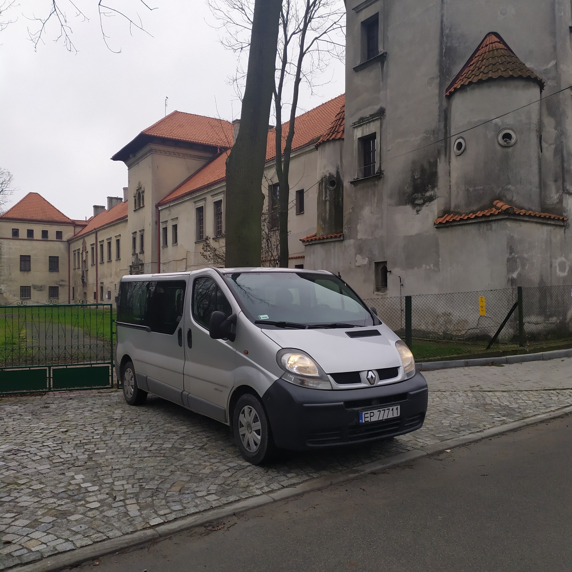 Srebrny van Renault Trafic zaparkowany na brukowanej nawierzchni przed zabytkowym budynkiem z czerwonym dachem i wieżyczką.