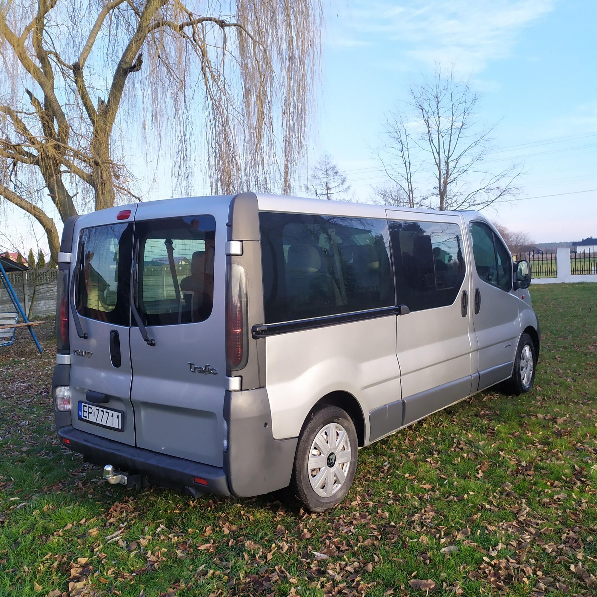 Srebrny Renault Trafic z hakiem holowniczym, zaparkowany na trawie pokrytej liśćmi, z widocznym numerem rejestracyjnym EP 77711, na tle drzew bez liści i błękitnego nieba.