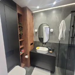 Nowoczesna łazienka z szarymi płytkami imitującymi marmur, czarnymi meblami, owalnym lustrem i białym zlewem nablatowym, oświetlona punktowymi lampami sufitowymi i taśmą LED.