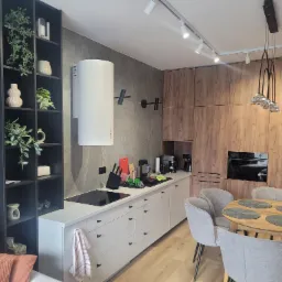 Nowoczesna kuchnia w odcieniach szarości i drewna, z wyspą kuchenną, okrągłym stołem z szarymi krzesłami, i czarnym regałem z dekoracyjnymi roślinami.