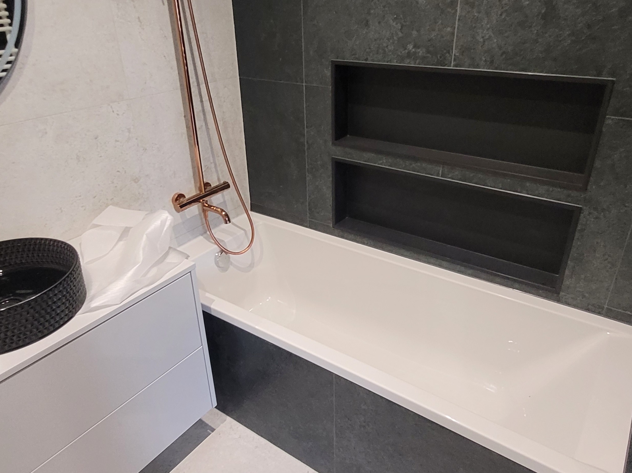 Nowoczesna łazienka z białą wanną, ciemnoszarą płytką i wnękami w ścianie, armatura w kolorze miedzi, umywalka nablatowa w kolorze czarnym.