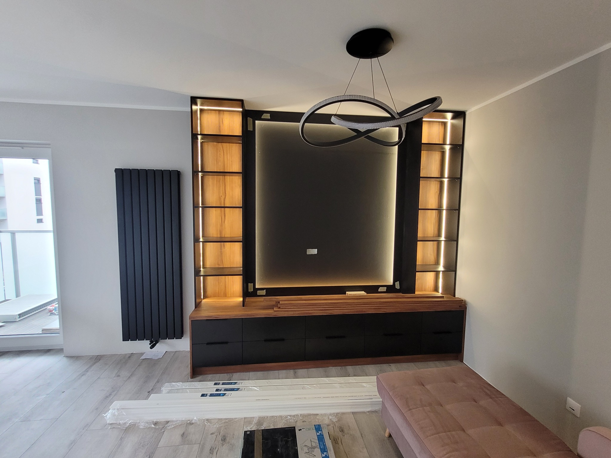 Nowoczesna zabudowa ściany z czarnym panelem TV, podświetlanymi półkami z drewnianym fornirem i czarnymi szafkami, uzupełniona o designerską lampę wiszącą i grzejnik panelowy.