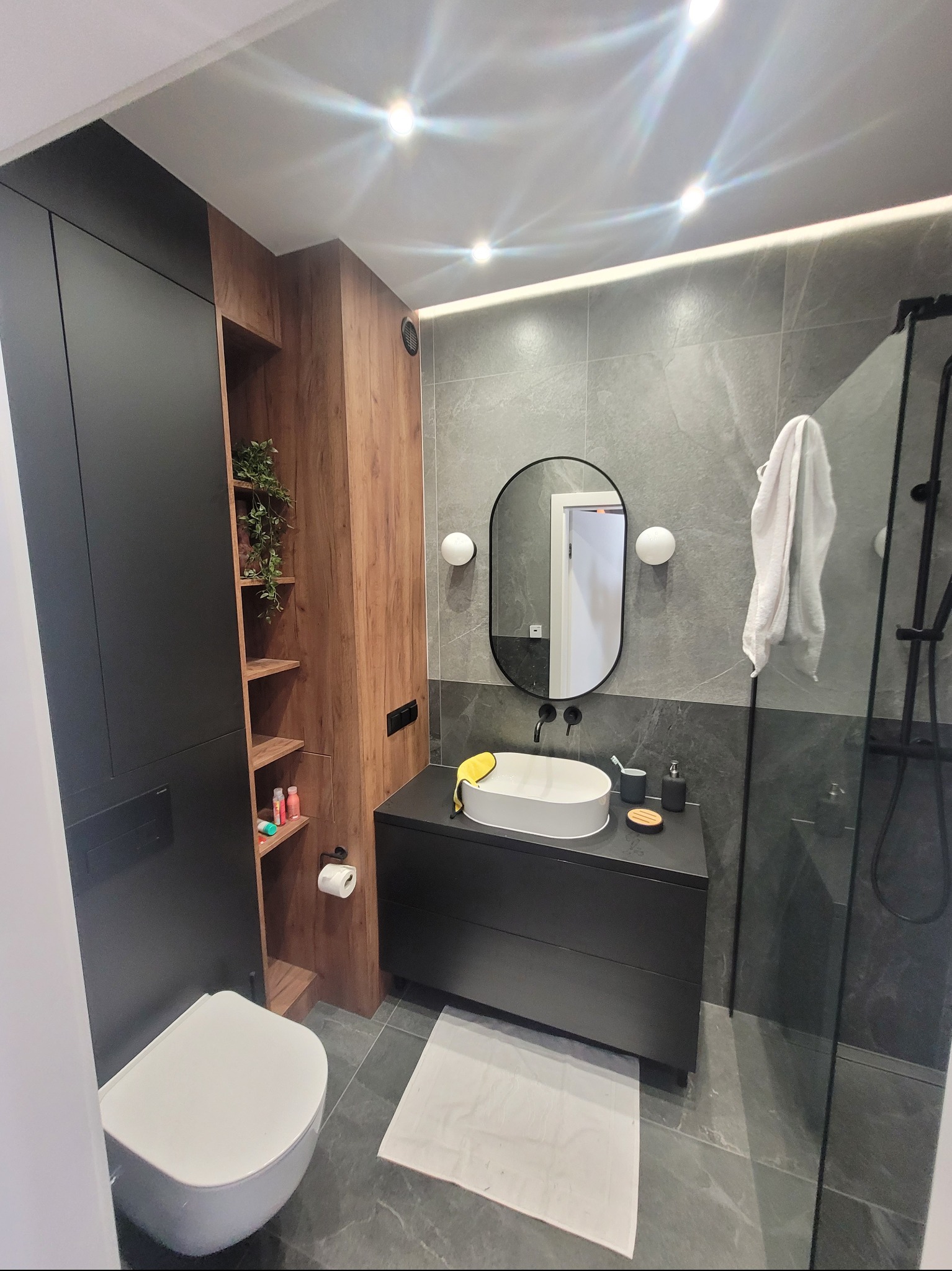 Nowoczesna łazienka z szarymi płytkami imitującymi marmur, czarnymi meblami, owalnym lustrem i białym zlewem nablatowym, oświetlona punktowymi lampami sufitowymi i taśmą LED.