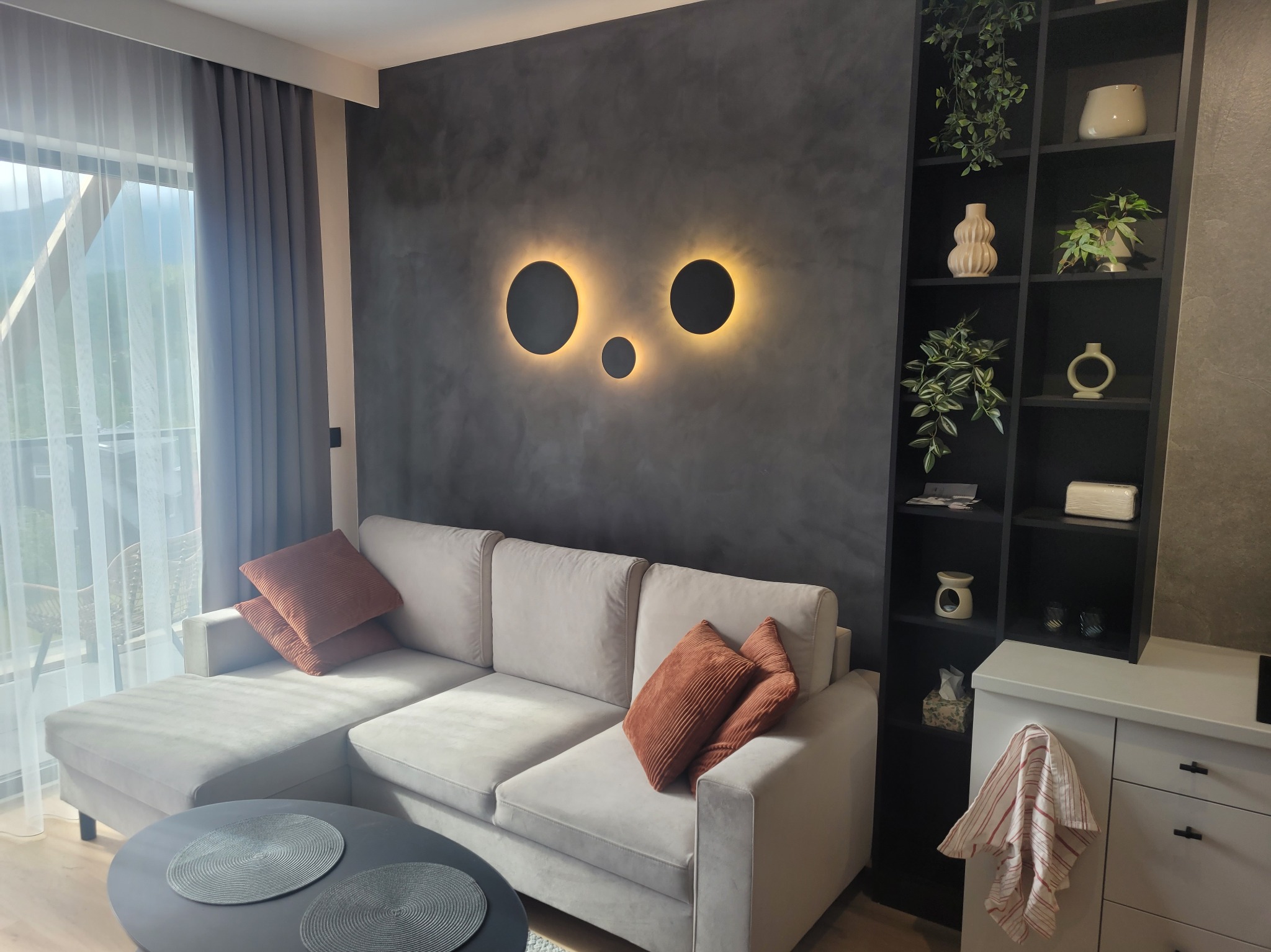 Jasnoszary narożnik z brązowymi poduszkami i okrągłym stolikiem na tle szarej ściany z trzema okrągłymi lampami, obok czarny regał z dekoracjami i biała komoda z zawieszoną ściereczką.