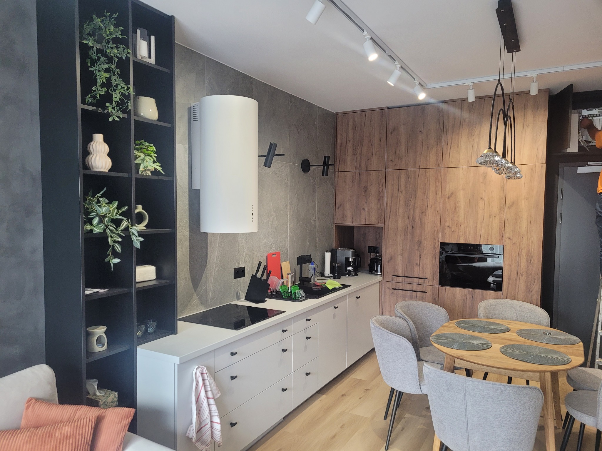 Nowoczesna kuchnia w odcieniach szarości i drewna, z wyspą kuchenną, okrągłym stołem z szarymi krzesłami, i czarnym regałem z dekoracyjnymi roślinami.