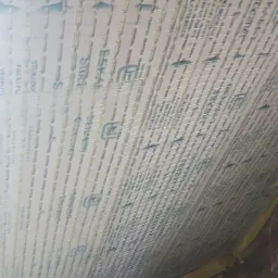 Montaż siatki z włókna szklanego na płycie izolacyjnej z nadrukowanym logo producenta, przygotowanie do prac wykończeniowych wewnątrz budynku, widoczne fragmenty drewnianej konstrukcji poddasza.