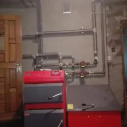 Czerwono-szary piec Tekla na paliwo stałe z podajnikiem, widoczne rury instalacji grzewczej i pompy obiegowe w kotłowni.