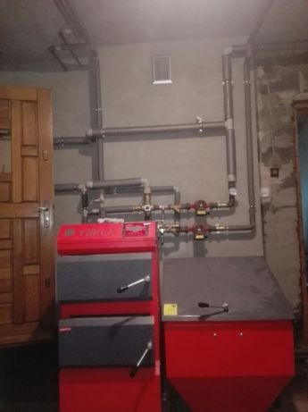 Czerwono-szary piec Tekla na paliwo stałe z podajnikiem, widoczne rury instalacji grzewczej i pompy obiegowe w kotłowni.