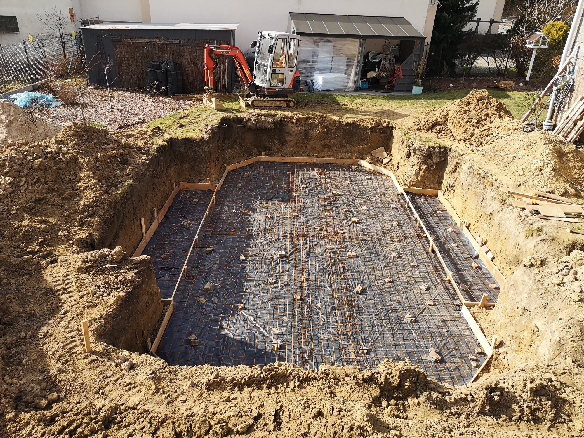 Wykop pod fundamenty z widocznym zbrojeniem siatką i deskowaniem, w tle minikoparka Kubota i budynek gospodarczy.