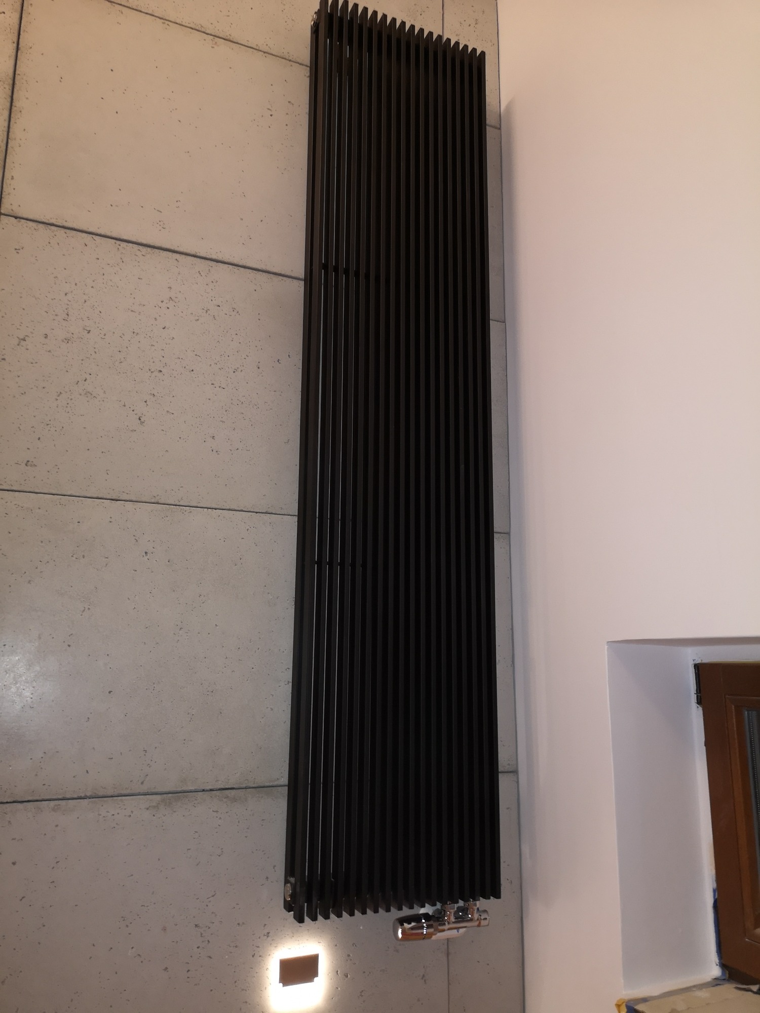 Wysoki, czarny grzejnik panelowy zamontowany na ścianie wyłożonej betonowymi płytami, z widocznymi chromowanymi zaworami i podświetleniem LED u dołu, obok fragmentu okna.